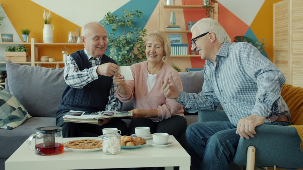 Old people friends are watching photos talking sitting at table with food in apartment enjoying communication together. Friendship and lifestyle concept.